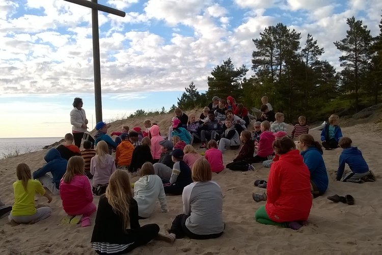 Barn som sitter i sanden vid stranden vända mot havet och ett träkors. En ledare håller andakt