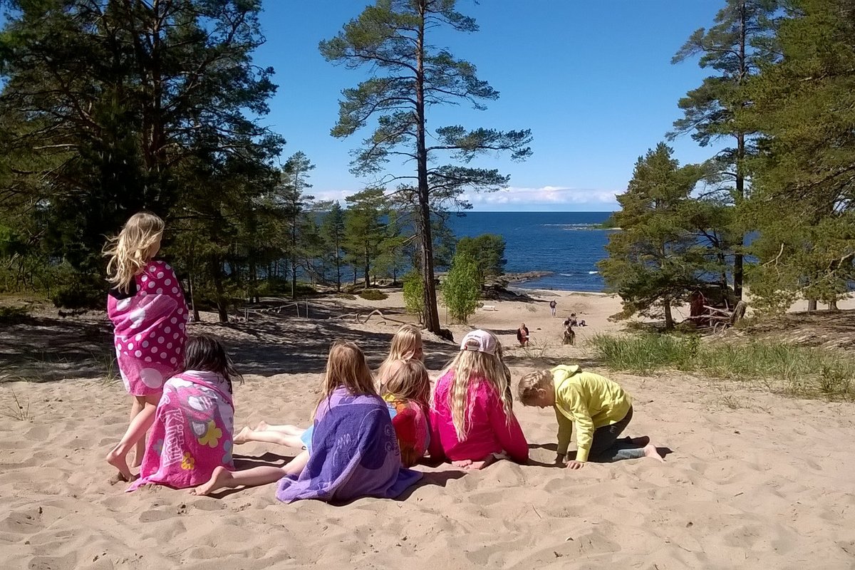 Barn insvepta i färggranna handdukar på sandstrand. Tallar och hav i bakgrunden
